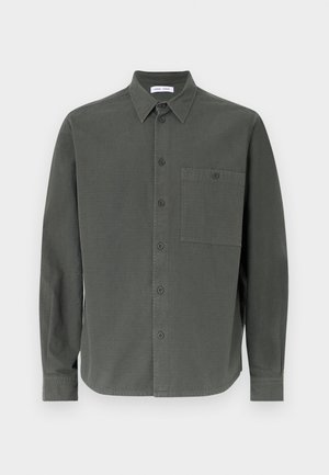 Tumši zaļš krekliņš ar pogām, izgatavots no kokvilnas, ar klasisku apkakli, garām piedurknēm un vienu krūšu kabatu ar pogu.