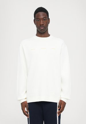 White crew neck sweatshirt made of cotton blend. Features gold text on the front. Loose fit with ribbed cuffs and hem. Worn with dark pants.