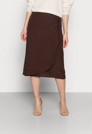 Vrouw draagt een donkerbruine wikkelrok tot op de knie, een witte trui en beige sandalen met blokhak en enkelbandje, staand tegen een effen achtergrond.