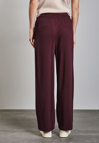 Pantalons larges bordeaux avec un waistband élastique, deux poches arrière et une texture lisse, présentés vue de dos.