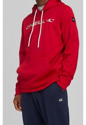 Person trägt einen roten O'Neill-Hoodie mit weißen Kordeln und marineblaue Jogginghose mit kleinem weißem Logo am linken Oberschenkel.