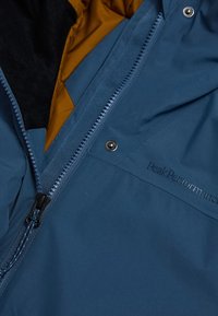 Blauwe waterdichte jas met een rits aan de voorkant, fleece-gevoerde kraag en contrasterende gele binnenkant met 'Peak Performance' branding.