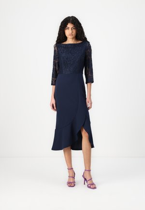 Robe bleu marine avec des manches en dentelle trois-quarts, corsage ajusté et ourlet évasé avec une fente latérale. Associée à des talons à lanières violets.