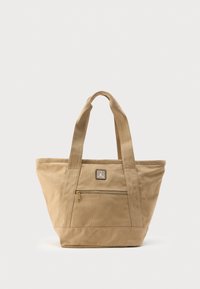Sac fourre-tout en toile beige avec deux bretelles, poche zippée à l'avant, et petit écusson carré avec logo au-dessus de la poche.