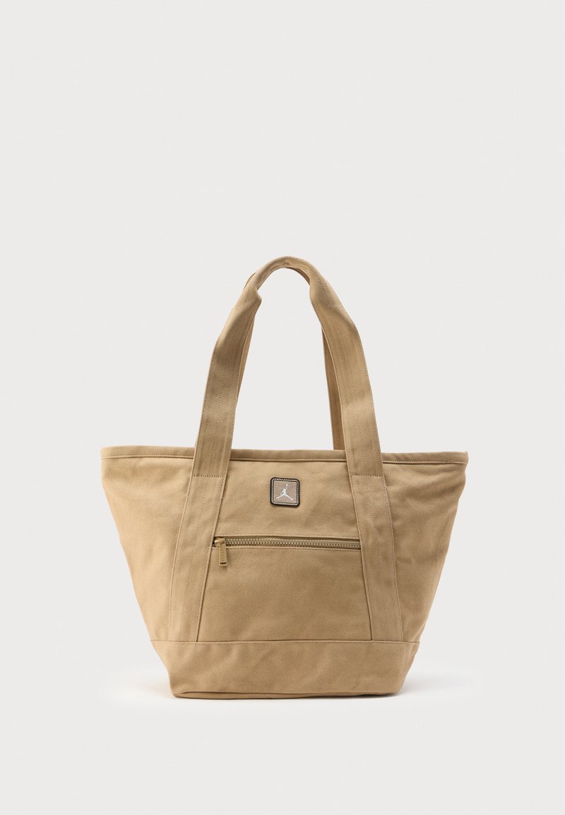 Sac fourre-tout en toile beige avec deux bretelles, poche zippée à l'avant, et petit écusson carré avec logo au-dessus de la poche.