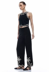 Svart crop top med vita broderade scalloped kanter, i kombination med vida svarta byxor med dekorativa palmträdsmönster vid fållen.