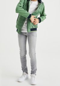 Grön vadderad jacka med dragkedja, marinblå detaljer, vit t-shirt, ljusgrå jeans och vita sneakers, som visar en avslappnad outfit.