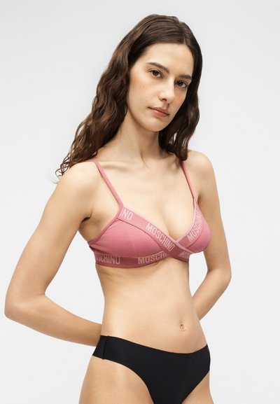 Γυναίκα με μακριά καστανά μαλλιά φοράει ροζ bralette Moschino και μαύρα εσώρουχα, στέκεται με τα χέρια πίσω από την πλάτη σε απλό φόντο.