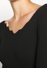 Haut en maille côtelée noire avec un décolleté en V festonné et des manches longues. Le tissu présente une texture avec une coupe ajustée.