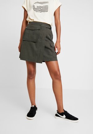Persona che indossa una gonna a portafoglio verde oliva con grandi tasche, una t-shirt bianca con stampa grafica e sneaker Nike nere, in piedi davanti a uno sfondo semplice.