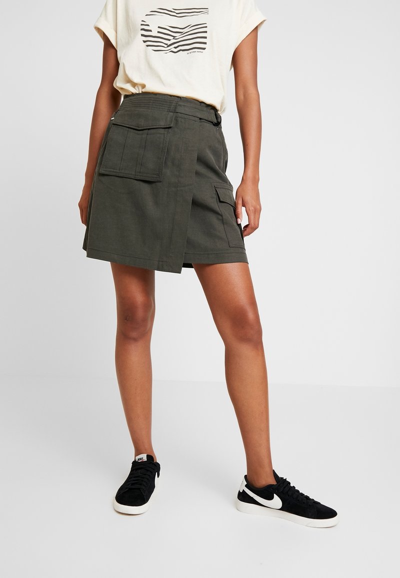 Personne portant une jupe portefeuille vert olive avec de grandes poches, un t-shirt blanc à motif graphique et des baskets noires Nike, debout devant un fond uni.