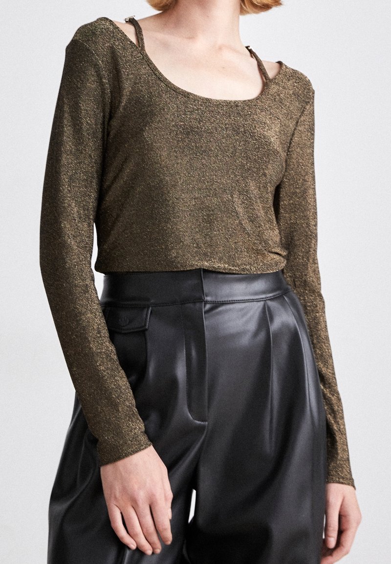 Bronze långärmad topp med rundad halsringning, utrustad med tunna axelband. Kombineras med högmidjade svarta läderbyxor.