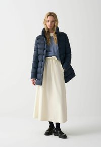 Marineblauwe pufferjack, lichtgewicht met een rechte pasvorm, over een lichtblauw sweater en een crèmekleurige enkellange rok. Zwarte schoenen met dikke zolen.