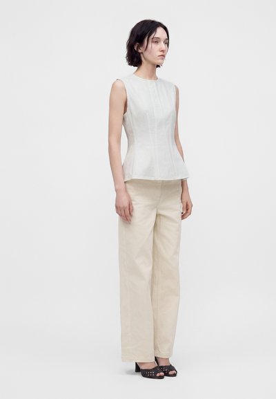 Femme portant un haut blanc ajusté sans manches, un pantalon beige à jambes larges et des chaussures noires à talons ouverts, debout devant un fond uni.
