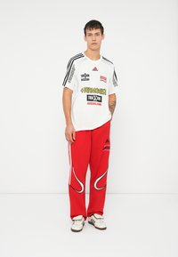 Balta Adidas T-krekls ar melniem un sarkaniem logo, apvienots ar sarkaniem sporta biksēm, kas aprīkotas ar baltām izliekta raksta detaļām un melno Adidas zīmolu.