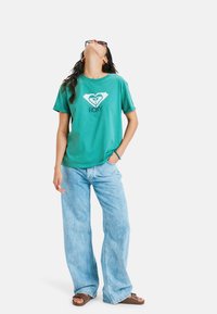 Grøn bomulds t-shirt med et hvidt logo design, parret med lyseblå brede jeans og brune sandaler. Simpelt og afslappet stil.