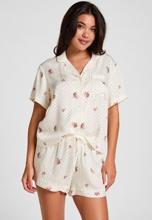Frau trägt cremefarbene Pyjamas mit kurzen Ärmeln, kleinen schwarzen Punkten und rosa Blumenmustern, knöpfbares Oberteil und Shorts mit Kordelzug, steht vor einfarbigem Hintergrund.
