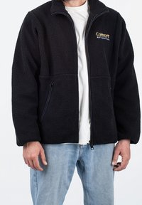 Czarna polarowa kurtka z zamkiem błyskawicznym z przodu, dwoma bocznymi kieszeniami oraz logo Carhartt na lewej stronie klatki piersiowej. Miękki, teksturowany materiał.