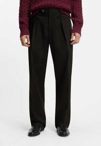 Pantaloni neri a pieghe con una vestibilità sartoriale, realizzati in tessuto morbido. Abbinati a scarpe nere senza lacci e a un maglione bordeaux a trama.