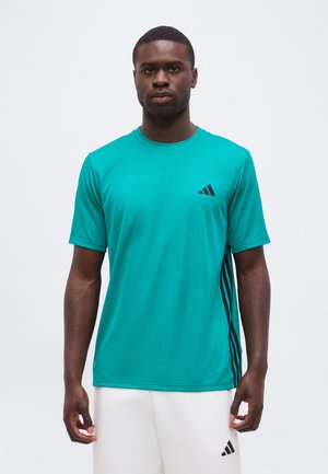Hombre de pie con una camiseta deportiva de manga corta color verde azulado con rayas negras y pantalones cortos blancos contra un fondo liso.
