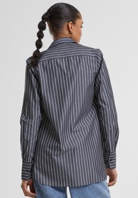 Donkergrijze blouse met witte verticale strepen, lange mouwen en een afgeronde zoom. Heeft een standaard kraag en een relaxed fit.