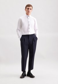 Man staat met zijn handen in zijn zakken, gekleed in een witte blouse met knopen, marineblauwe geplooide broek, zwarte sokken en zwarte nette schoenen.