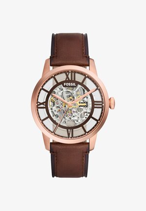 Montre automatique homme Fossil avec bracelet en cuir marron, boîtier en or rose, cadran squelette montrant des rouages complexes et des index horaires en chiffres romains.