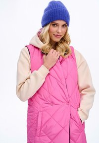 Różowa pikowana kamizelka z kapturem i kremowymi rękawami, z przodu zapinana na napy i z bocznymi kieszeniami, połączona z niebieską dzianinową czapką.