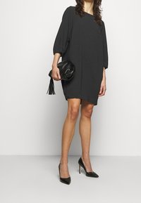 Vestido negro de corte holgado con mangas tres cuartos, combinado con un bolso de mano negro con un acento de borlas y tacones negros puntiagudos.
