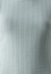 Top en tricot bleu clair présentant une texture côtelée verticale avec de petits motifs décoratifs. Vue rapprochée mettant en valeur les détails du matériau et du design.