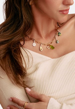 Femme portant un collier chaîne en or avec des breloques variées, comprenant une fleur blanche, un avocat, un ourson en gomme vert et une tortue, sur un top côtelé crème.