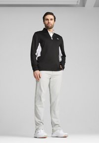 Schwarzer Pullover mit langen Ärmeln, weißen Seitenpaneelen, ¼-Reißverschluss und Logo; kombiniert mit hellgrauen Hosen und weißen Sportschuhen.
