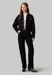 Chaqueta de mezclilla negra corta con botones plateados y dos bolsillos frontales, combinada con pantalones vaqueros negros de cintura alta y botines de tacón negro.