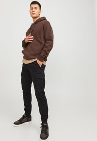 Brun hoodie med dragsko, beige lagertröja under, svarta cargobyxor med sidofickor och svarta sneakers med ljusa accenter.