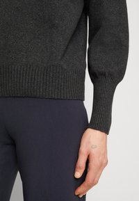 Pull en maille gris foncé avec poignets et ourlet côtelés. Le tissu semble doux, avec une texture légèrement marquée. La main repose sur un pantalon sombre.