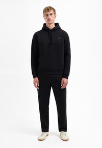 Schwarzer Fleece-Hoodie mit Kordelzügen, gerippten Bündchen und kleinem Logo. Kombiniert mit passenden schwarzen Jogginghosen und weißen Sneakern.