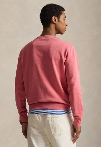 Άνδρας που κοιτάζει πλαγίως, φοράει μακρυμάνικο φούτερ κοραλί ροζ, ριγέ μπλε και λευκό πουκάμισο από μέσα και εκρού παντελόνι, με ουδέτερο φόντο.