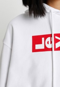 Felpa bianca in cotone con un grande logo rettangolare rosso con testo bianco sulla parte anteriore. Vestibilità ampia con cappuccio e cordoncino.