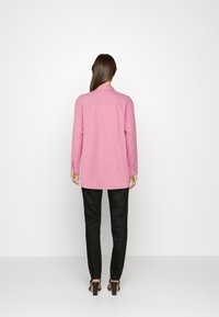 Camicia rosa a maniche lunghe con colletto, polsini abbottonati e taglio dritto. Abbinata a pantaloni neri aderenti e scarpe con tacco.