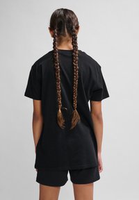 Camiseta de algodón negra con un corte holgado, combinada con pantalones cortos negros. Cabello largo y trenzado cae por la espalda. Fondo gris neutro.