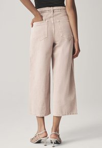 Nő világos rózsaszín, széles szárú, rövid farmert visel, bézs színű magassarkú szandállal, hátulról látható, egyik kezét a zsebében tartva.