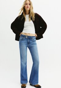 Temno rjava kratka jakna, bela prilega punca ter svetlo modri razširjeni jeans. Jeans imajo sprednje žepe in svetlo oprano tkanino. Rjave gležnjače.