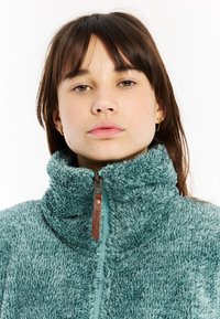 Mlada žena smešnih smešnih ima smešnu smešnu majicu u boji teal s visokim ovratnikom na beloj pozadini.