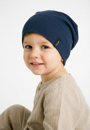 Young child wearing a beige outfit and navy blue beanie, smiling, sitting against a plain white background