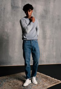 Jasnoszary sweter, niebieskie dżinsy i białe trampki. Model stoi na wzorzystym dywanie przed szarym murem, z teksturowaną powierzchnią i miękkim oświetleniem.