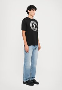 Joven de pie de perfil, vistiendo una camiseta gráfica negra, jeans azul claro y zapatos negros, contra un fondo blanco liso.