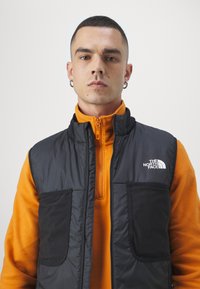 Black insulated vest with two front pockets, over an orange fleece top. The North Face logo is visible on the vest's upper left side.