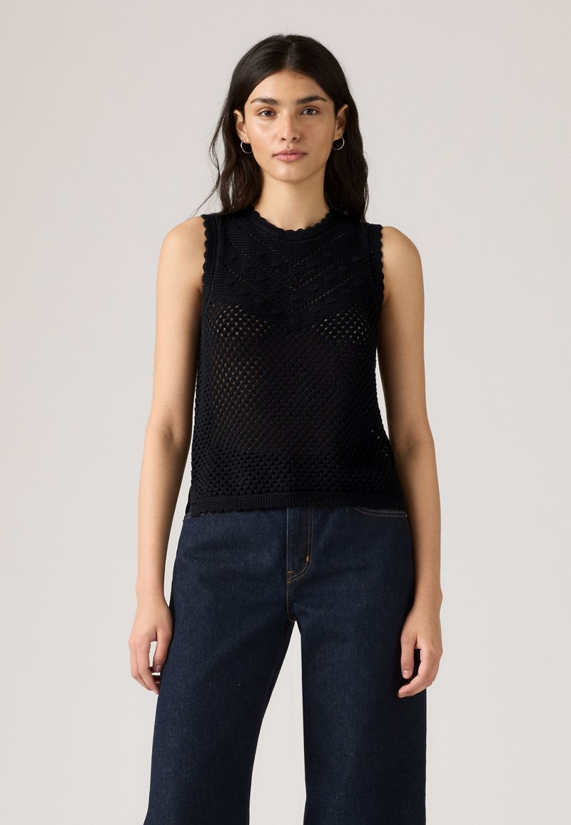 Młoda kobieta w bezrękawnikowej czarnej siatkowej bluzce i ciemnoniebieskich szerokich spodniach, stojąca na jednolitym jasnym tle.