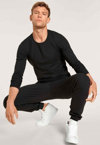 Chemise noire à manches longues et joggers noirs avec des accents rouges sur les côtés, associés à des baskets blanches montantes, en position accroupie.