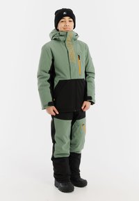 Groene en zwarte waterdichte jas en broek set met oranje accenten, voorzien van ritszakken en een hoge kraag voor warmte en bescherming.